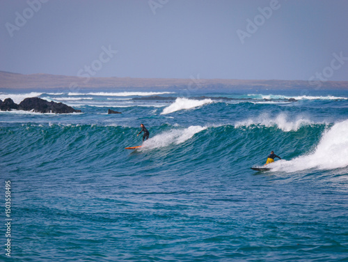 Photography Two surfers catch a clean wave on a sunny day near a rocky, volcanic coastline