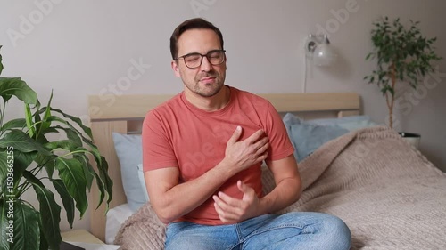 A man sits on a bed, suddenly clutching his chest in pain. His face expresses fear and anxiety. Heart attack, infarction. Concept: cardiology, health.