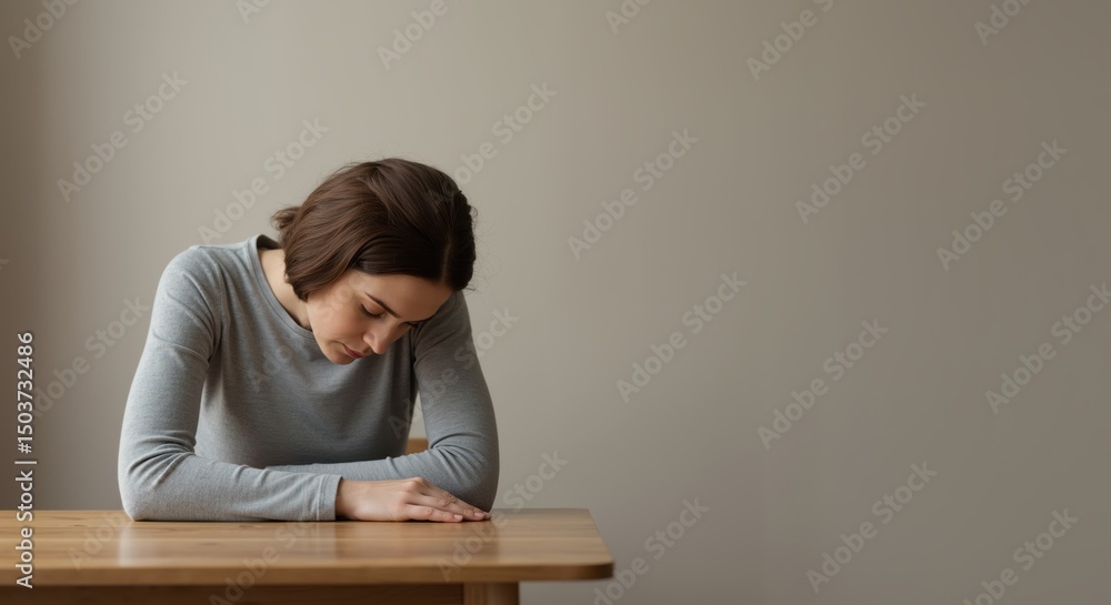 Young woman sitting at table with head down in neutral room Young woman sitting at table with head down in neutral room