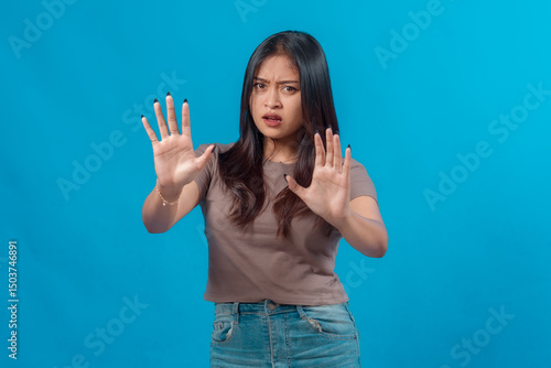 Young Asian woman looking concerned and raising both hands in a stop gesture, signaling rejection or warning, while standing in casual outfit against a plain blue background.