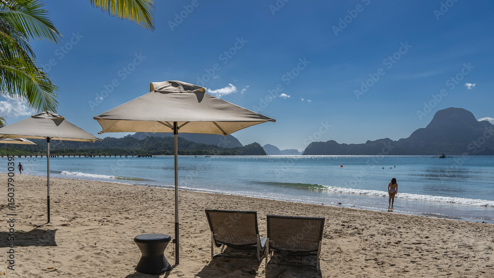 Fototapeta premium A sunny day, a tropical beach. Two sun loungers under sun umbrellas. The waves of the calm turquoise ocean are foaming. Silhouettes of people. Mountains on the horizon.Palm leaves against the blue sky