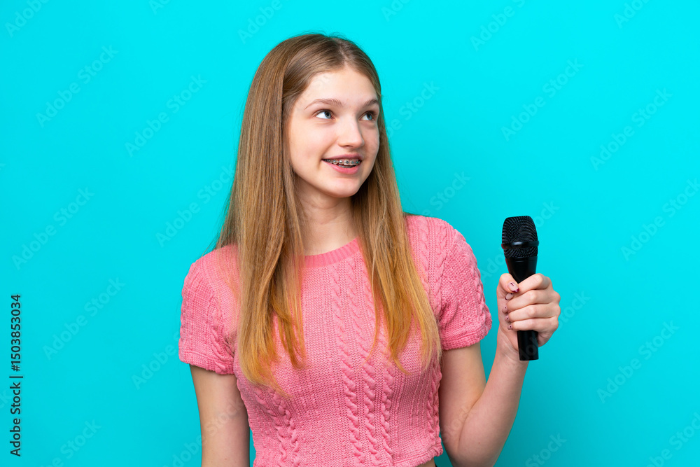 Singer Russian girl picking up a microphone isolated on blue background looking up while smiling Singer Russian girl picking up a microphone isolated on blue background looking up while smiling