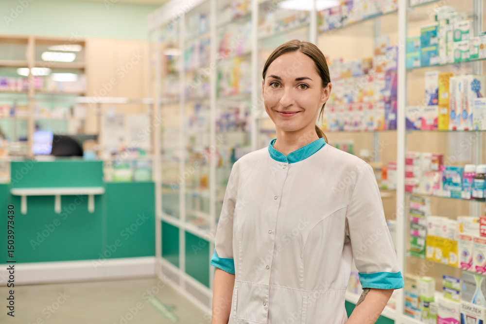 Obraz premium Smiling young female pharmacist with brown hair stands confidently in well-lit pharmacy. Shelves filled with colorful medicines in background create vibrant ambiance