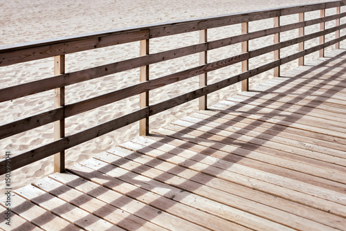 Sunlit wooden boardwalk stretches along serene sandy beach. Shadows from railing create pattern on wooden planks, evoking peaceful and tranquil ambiance