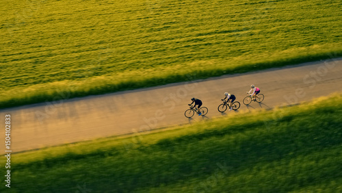 An aerial view captures three cyclists racing down a scenic country road, surrounded by lush green fields. The dynamic perspective emphasizes their speed and the exhilaration of road cycling.