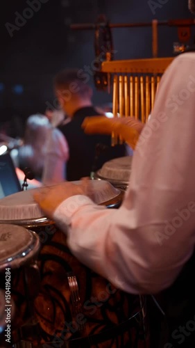 Close-up of a percussionist playing conga drums during a live music performance, with warm stage lighting and a blurred background of musicians.