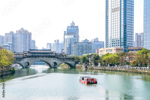 Scenic landscape shot of tour boat on the Jinjiang River about to cross beneath the iconic ancient Anshun Bridge in Chengdu, modern buildings in the backgroung