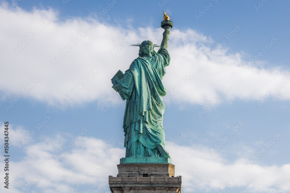 Obraz premium Rear view of the Statue of Liberty with torch raised, seen against a partly cloudy blue sky in New York City. Iconic symbol of freedom and democracy in the United States.