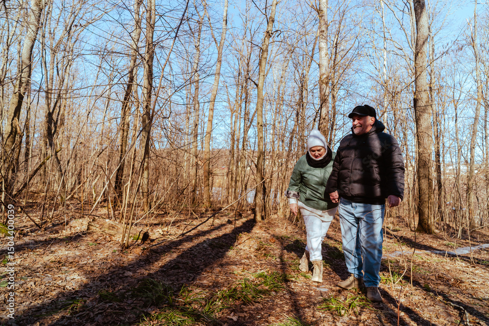 Obraz premium An elderly couple, a man and a woman, walk through a spring forest, enjoying the beauty of nature. Sunlight filters through fresh green leaves, creating a serene and romantic atmosphere