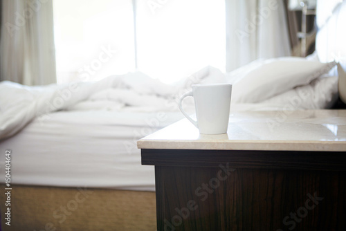 white cup on the bedside table near empty white bed sheets, seen through large window.