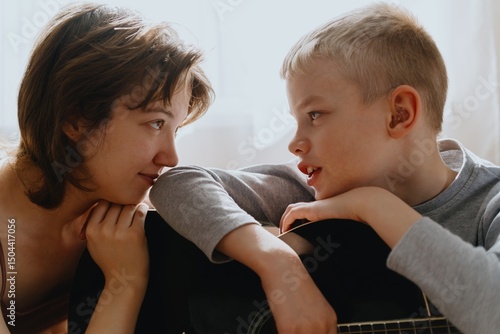 Mother and son playing guitar together, family love. The child wears a hearing aid, highlighting inclusivity and the universal language of music. Authentic, unedited photo captures emotions