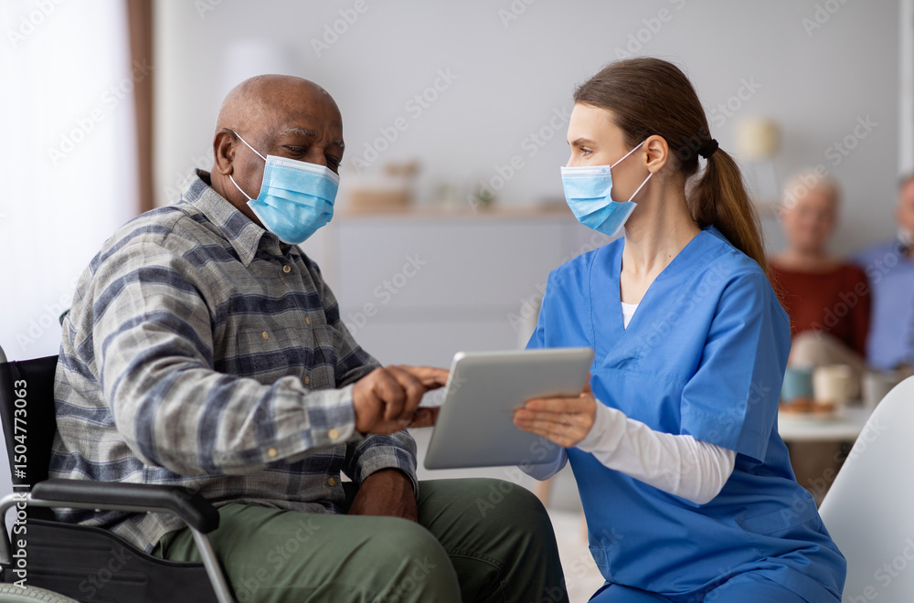 Obraz premium Female nurse helping black senior man in weheelchair using digital tablet, elderly african american male patient surfing on Internet at nursing home, wearing protective face masks, copy space