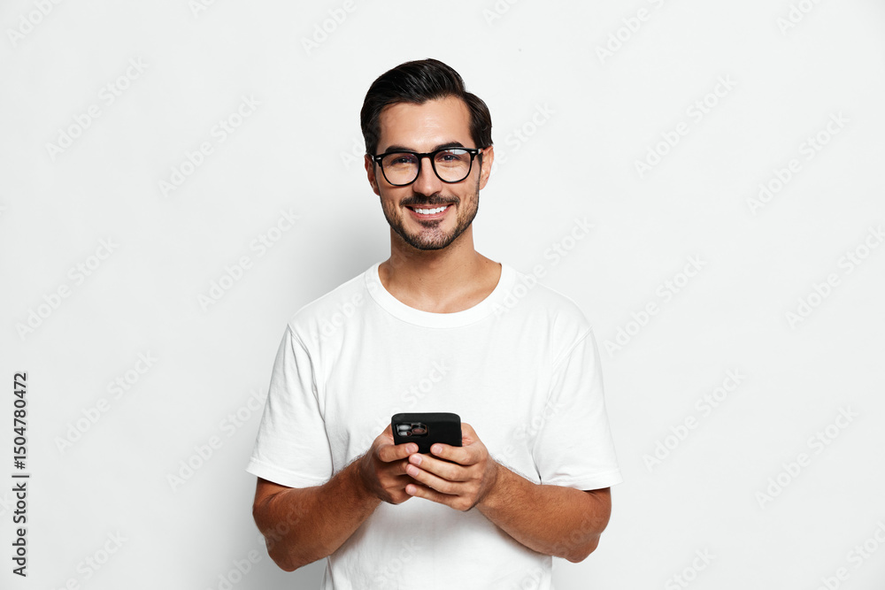 Fototapeta premium Smiling man wearing glasses holds smartphone in hand, staring at camera in casual white t-shirt against plain solid background. Lifestyle concept.