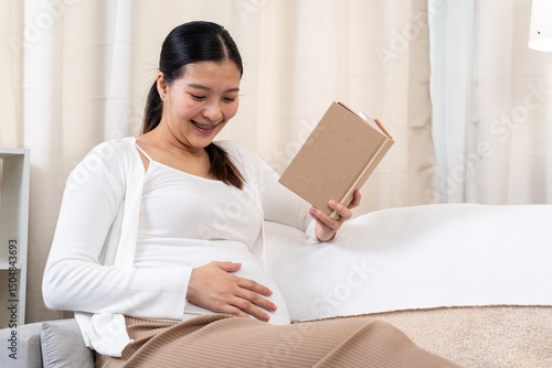 Pregnant Asian woman smiling while reading book and touching belly. Wearing white cardigan and brown pants. Cozy home setting expresses joy, connection, maternal care, and learning for motherhood. Free Download Pregnant Asian woman smiling while reading book and touching belly. Wearing white cardigan and brown pants. Cozy home setting expresses joy, connection, maternal care, and learning for motherhood. - GFXLoad
