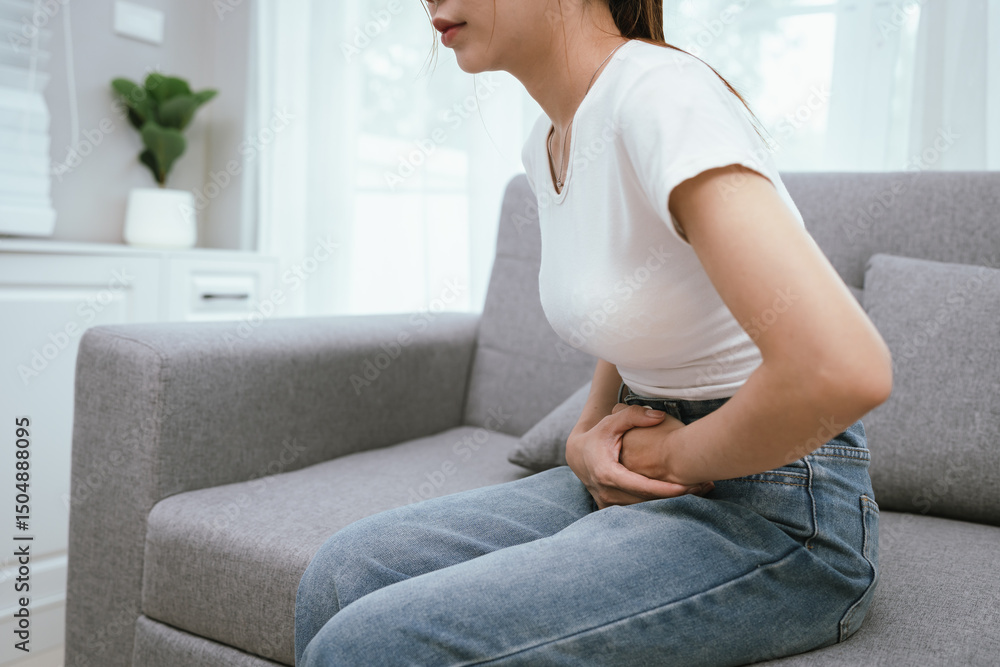Young adult woman sitting on a sofa, holding her abdomen in discomfort, experiencing menstrual cramps or digestive pain, looking visibly uncomfortable. Young adult woman sitting on a sofa, holding her abdomen in discomfort, experiencing menstrual cramps or digestive pain, looking visibly uncomfortable.