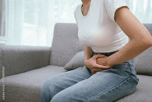 Wall Mural Young adult woman sitting on a sofa, holding her abdomen in discomfort, experiencing menstrual cramps or digestive pain, looking visibly uncomfortable