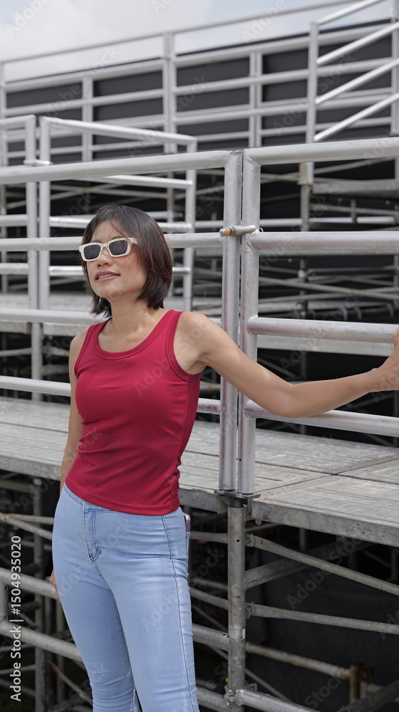 Fototapeta premium Pretty Asian woman stands on outdoor metal stands, showcasing modern casual fashion with a bold red sleeveless top and light blue jeans