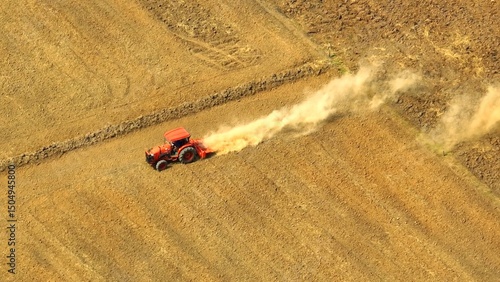 Across the sun-scorched land, a tractor hauls a cultivator, leaving a trail of swirling dust in its wake, embodying the enduring spirit of farm labor amidst harsh conditions.
