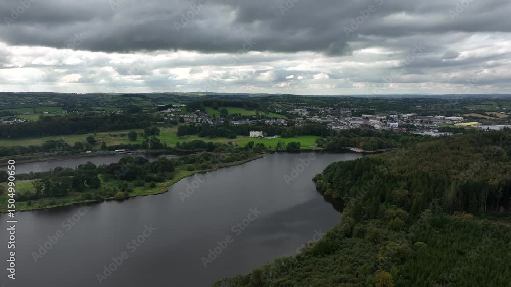 Lough Muckno, County Monaghan, Ireland, September 2022. Drone high angle descends low over grey waters approach old castle on opposing lake shore hill side under overcast sky. Lough Muckno, County Monaghan, Ireland, September 2022. Drone high angle descends low over grey waters approach old castle on opposing lake shore hill side under overcast sky.