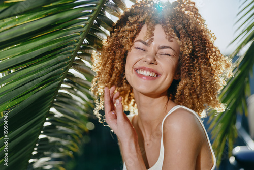 Φωτογραφία Smiling woman with curly hair enjoys sunny tropical outdoors, surrounded by green palm leaves, radiating happiness and natural beauty in warm daylight