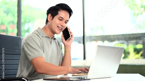 Confident Asian man talking on smartphone and working on laptop at office desk.