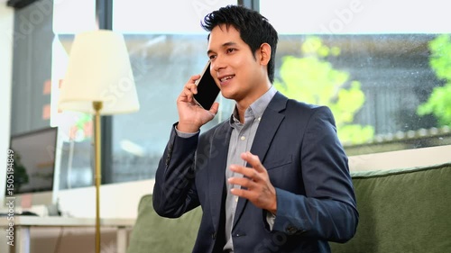 Confident Asian man talking on smartphone and working on laptop at office desk.