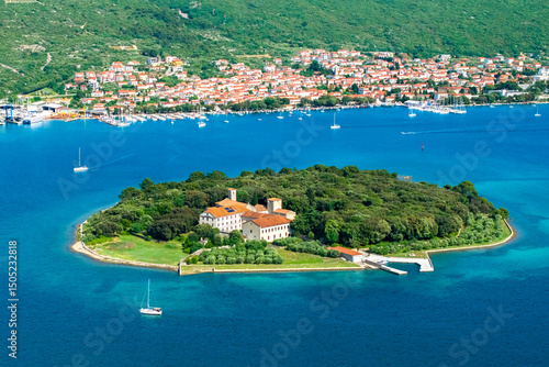 A wide aerial view showcasing Košljun Island with its monastery, surrounded by calm blue waters and the coastal town of Punat in the background. Sailboats dot the sea