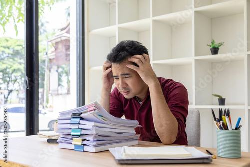 A man in a red shirt is sitting at a desk with a pile of papers in front of him. He is stressed and overwhelmed by the amount of work he has to do
