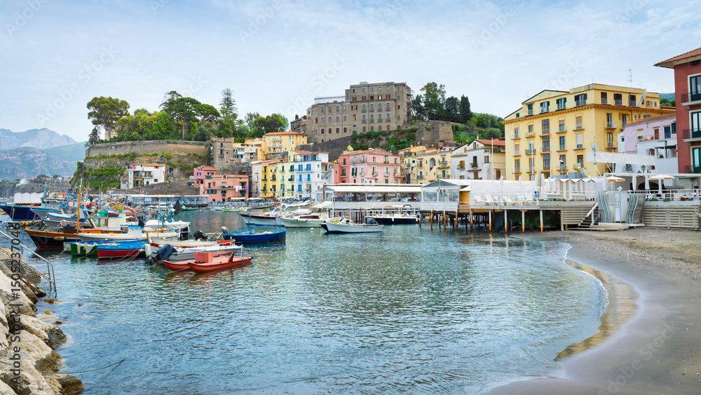Fototapeta premium Marina Grande, charming harbor in Sorrento, Italy. Colorful buildings, boats docked in harbor on calm sea water. Hills with lush green trees, cloudy sky create scenic European landscape