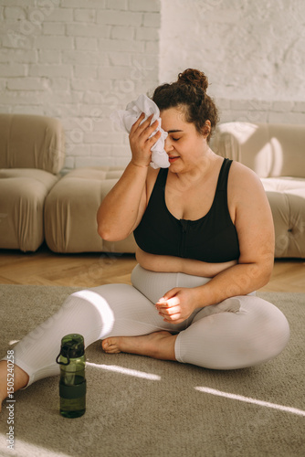 Fotografie A tired young curly-haired plus-size woman sits on the floor after a workout at home, drinks water and wipes sweat with a towel