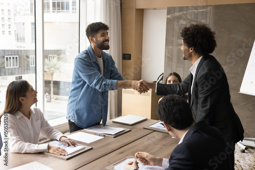 Negotiation success. Glad excited millennial male partner investor shake hand of business associate company owner on round table meeting with multiethnic professionals grateful for good proposal