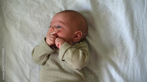Baby with blue eyes in olive overalls lies on back. Close-up. Child sucks finger on hand. First year of life and hygiene of child. Newborn - happiness for family. New worries and style life.