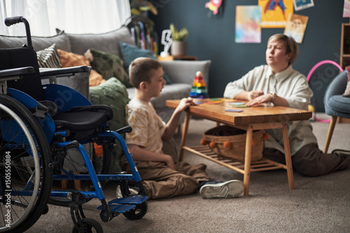 Canvas Print Young boy with disability is playing cards with a woman at a wooden table in a c