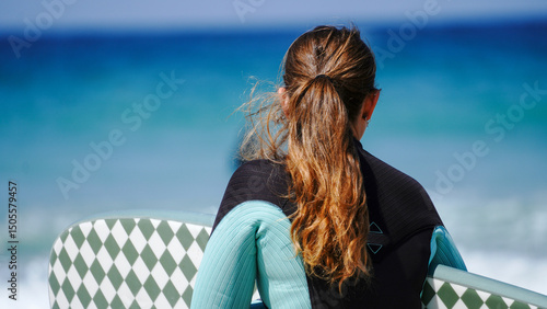 Fotografie A woman wearing a wetsuit holding a patterned surfboard looks out towards the ocean
