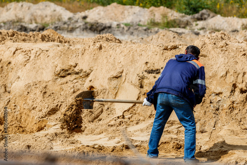 A man in a blue jacket and jeans diligently works with a shovel, digging sand at a construction site.
