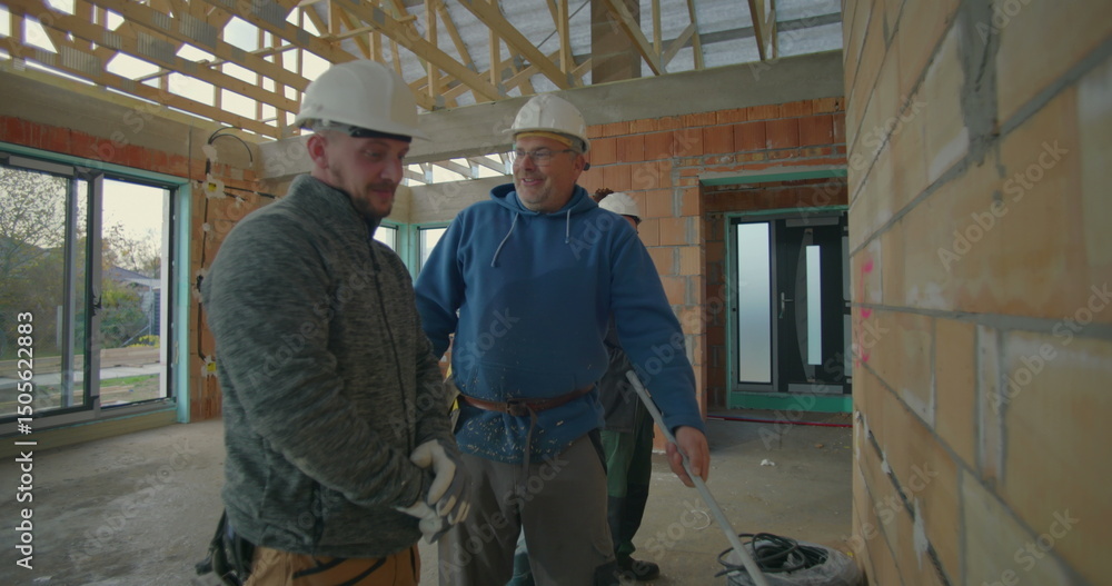 Construction workers discussing plans on site, three workers in hard hats and safety gloves reviewing blueprints in unfinished building interior Construction workers discussing plans on site, three workers in hard hats and safety gloves reviewing blueprints in unfinished building interior