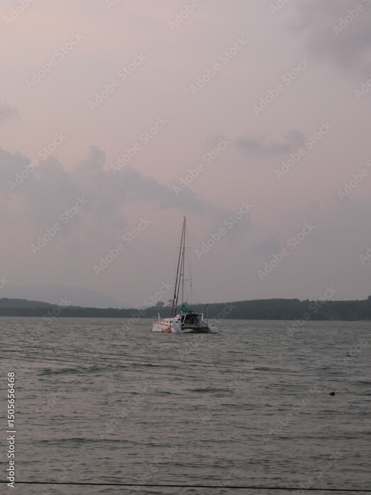 Obraz premium Serene view of a sailboat floating on calm waters at sunset, with soft pastel skies and distant forested hills in the background. Peaceful and dreamy seascape perfect for travel and nature themes.