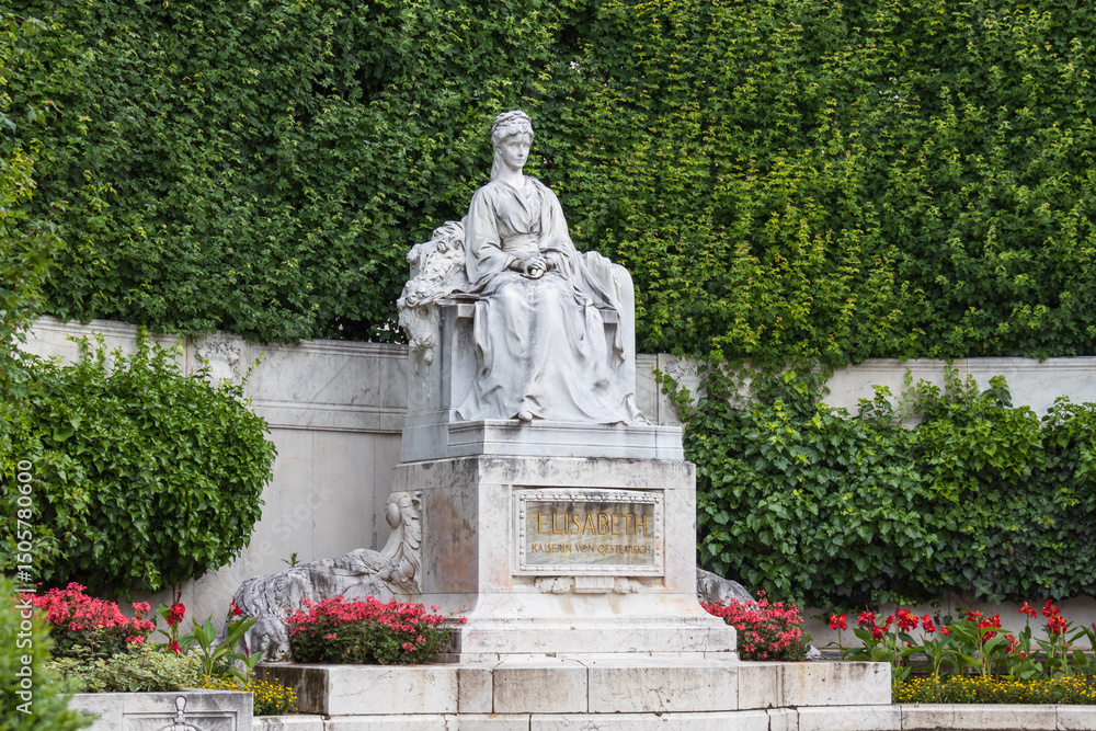 Fototapeta premium Marble statue of Empress Elisabeth of Austria (Sisi) seated in a serene garden setting in Volksgarten, Vienna. Surrounded by lush greenery and blooming flowers.