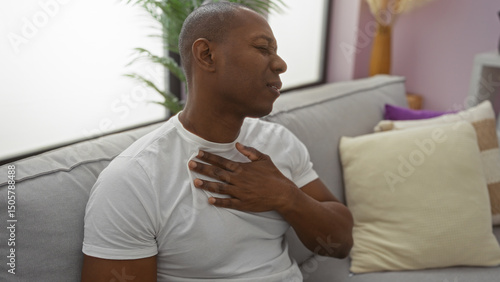 Foto Young man experiencing discomfort sitting on sofa in living room, showing expression of pain, hand on chest, inside modern apartment with cushions and plants