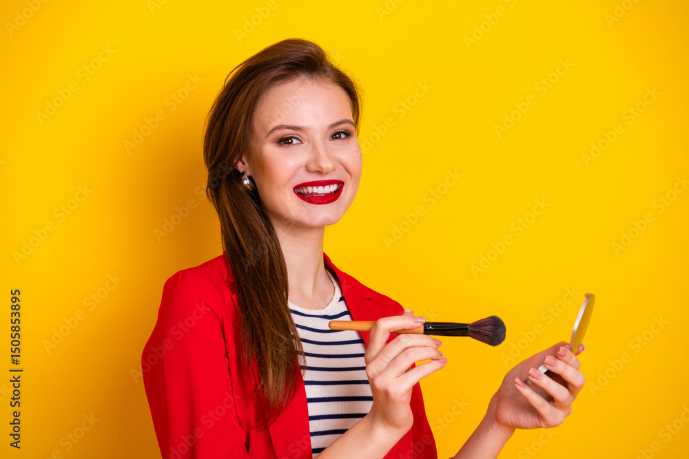 Young woman in a red jacket smiling enthusiastically while applying makeup, posing against a vibrant yellow background Young woman in a red jacket smiling enthusiastically while applying makeup, posing against a vibrant yellow background