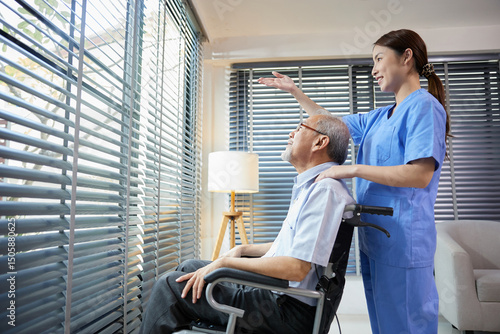 nurse or caregiver standing beside elderly man sitting on wheelchair and looking outside at home