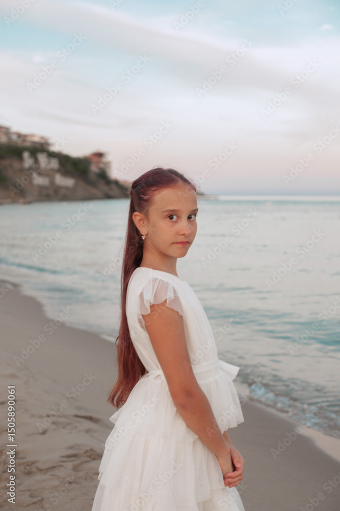 Side profile of a young girl in a white dress standing on the beach at sunset. Calm waves, pastel sky, and coastal cliffs in the background create a serene and natural atmosphere. Side profile of a young girl in a white dress standing on the beach at sunset. Calm waves, pastel sky, and coastal cliffs in the background create a serene and natural atmosphere.