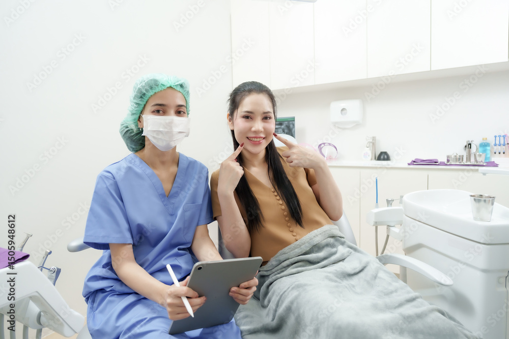 Fototapeta premium Female patient smiling and pointing at cheeks while sitting next to Asian dental assistant in blue uniform, both posing happily together in examination room after consultation session in dental clinic