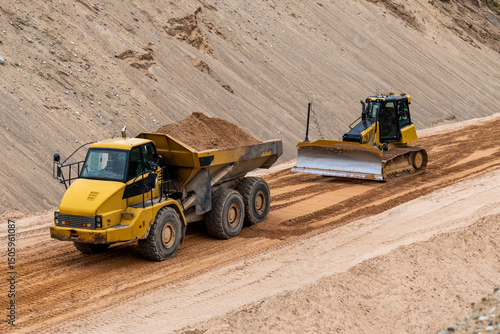 Heavy construction vehicles working on sandy site, dump truck and bulldozer leveling ground at quarry or infrastructure construction zone
