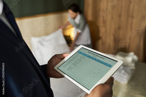 Business person managing hotel staff using tablet technology for housekeeping operations in hotel room setting while focusing on digital data and lists of duties