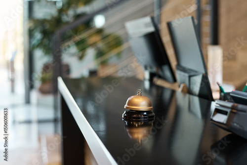 Gold bell on modern hotel reception desk reflecting ambient light, creating welcoming atmosphere. Computers in background hint at efficient check-in process