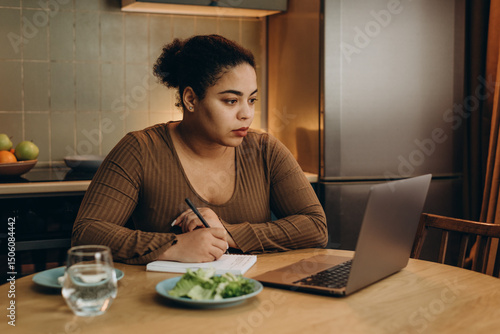 African American woman makes notes listening to webinar via laptop at table in kitchen, eating disorder concept