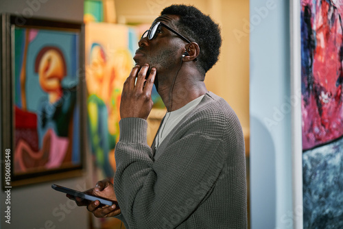 Man with headphones looking at painting in art gallery while holding phone. Engaging in thoughtful contemplation while surrounded by vibrant artwork in gallery