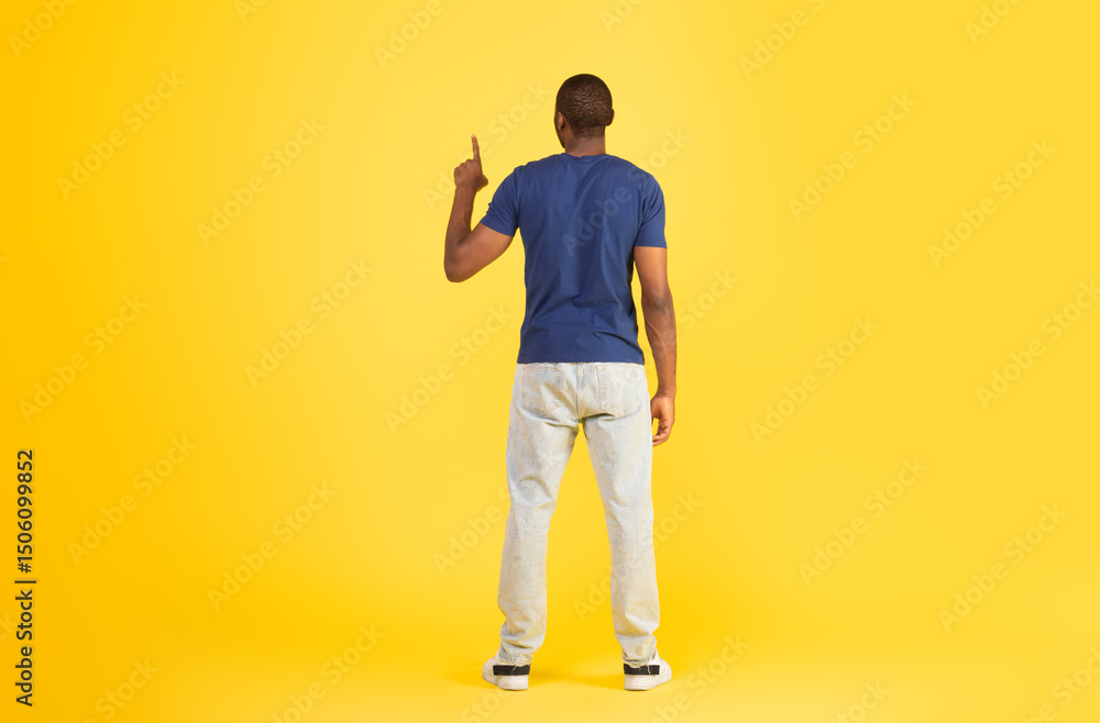 Fototapeta premium Back View Of African American Man Pointing Finger Up Pushing Invisible Button Or Touchscreen Standing Over Yellow Studio Background. Look Upward. Full Length Shot