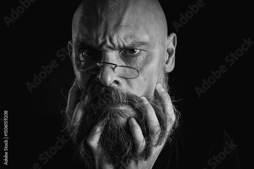 Εκτύπωση καμβά Black and white portrait, serious bald man with beard and glasses looking intently at the camera, holding beard in hands near face